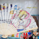 A folding hand fan with printed anime artwork and text, photographed indoors against a softly blurred background with colorful objects visible below.