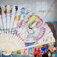 A folding hand fan with printed anime artwork and text, photographed indoors against a softly blurred background with colorful objects visible below.