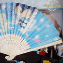 A folding hand fan with a light blue printed fabric featuring anime-style characters, stars, and pastel illustrations, attached to cream-colored wooden ribs. The fan is shown partially open against a cluttered indoor background with a watermark in the top right corner.
