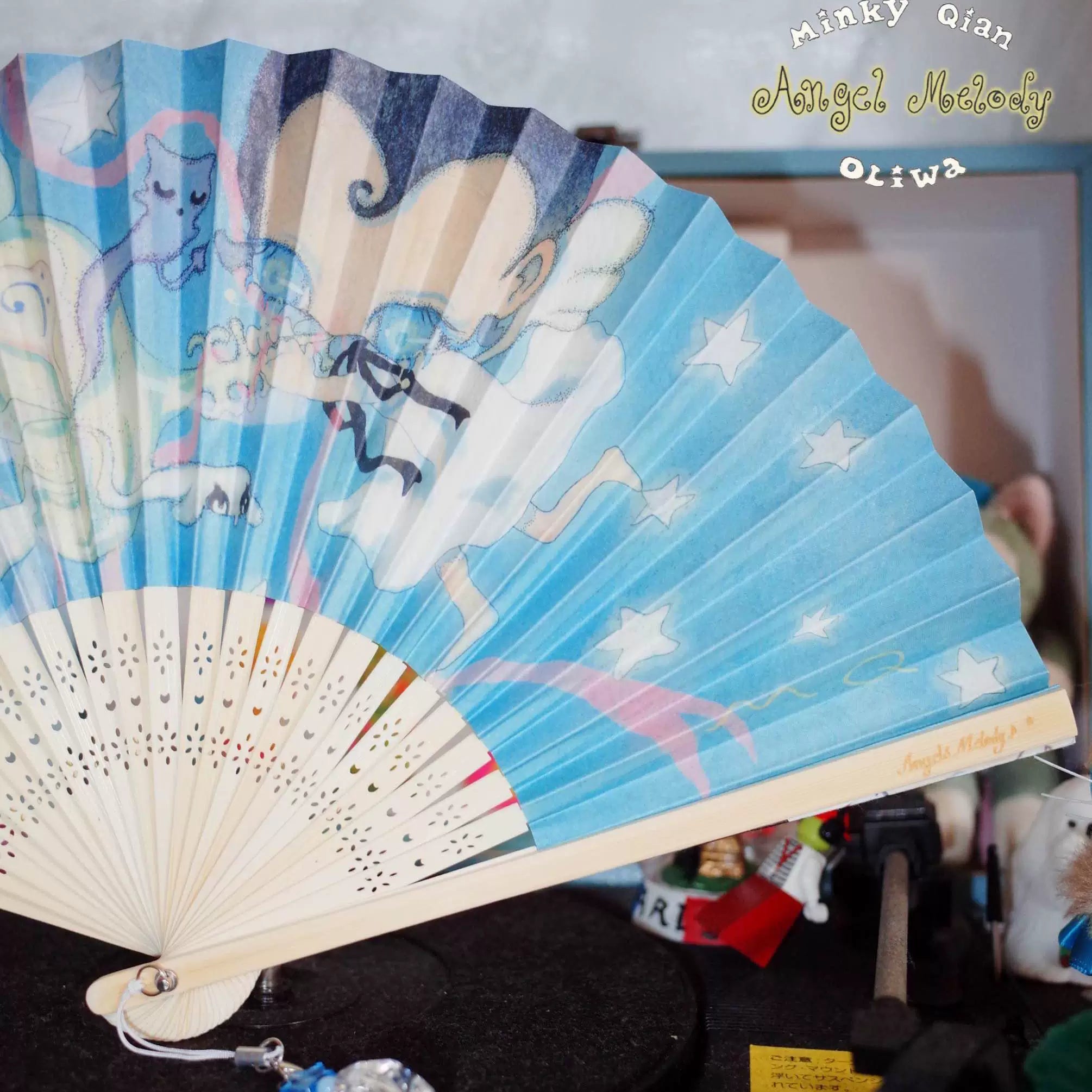 A folding hand fan with a light blue printed fabric featuring anime-style characters, stars, and pastel illustrations, attached to cream-colored wooden ribs. The fan is shown partially open against a cluttered indoor background with a watermark in the top right corner.
