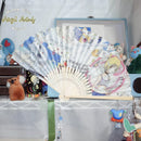 A folding hand fan with printed anime-style artwork and musical notes, displayed open on a shelf in a decorated room setting. The fan features characters and illustrations with pastel blue, pink, yellow, and white tones.
